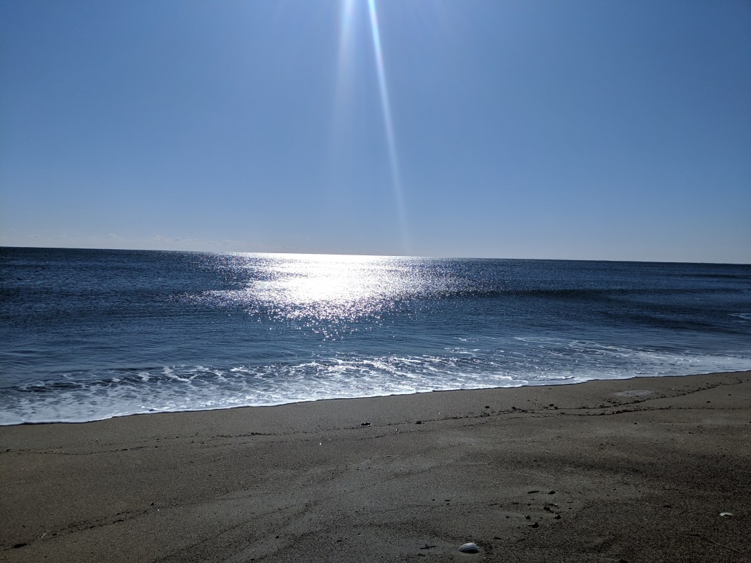 Sunlight reflecting across the surface of the ocean with a beach in the foreground, blue sky above.
