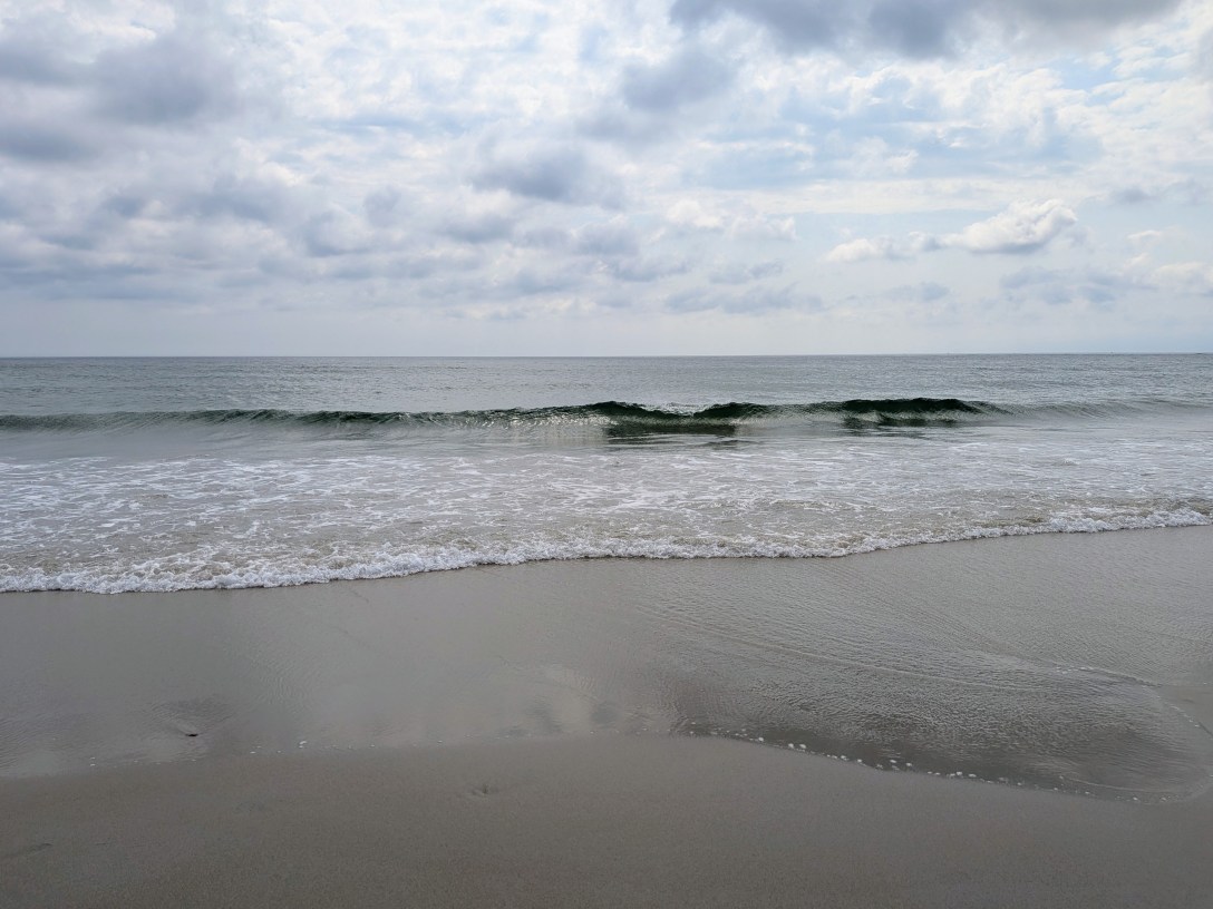 Calm grey ocean under a soft cloudy sky as a single wave rolls toward wet, reflective sand at Hampton Beach.