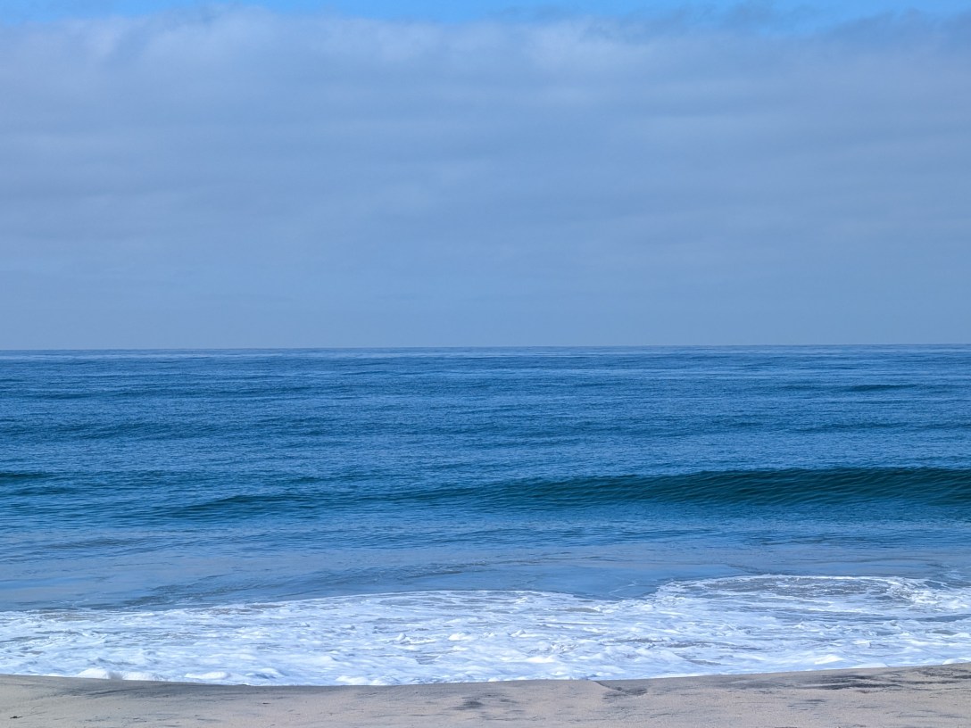 A single ocean swell rising toward shore before it breaks, under a soft hazy sky in cool blue tones.