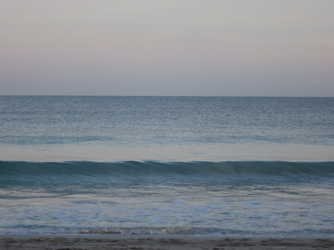 A soft, hazy seascape at dawn where sea and sky blur at the horizon, with a single gentle wave rising in the foreground.