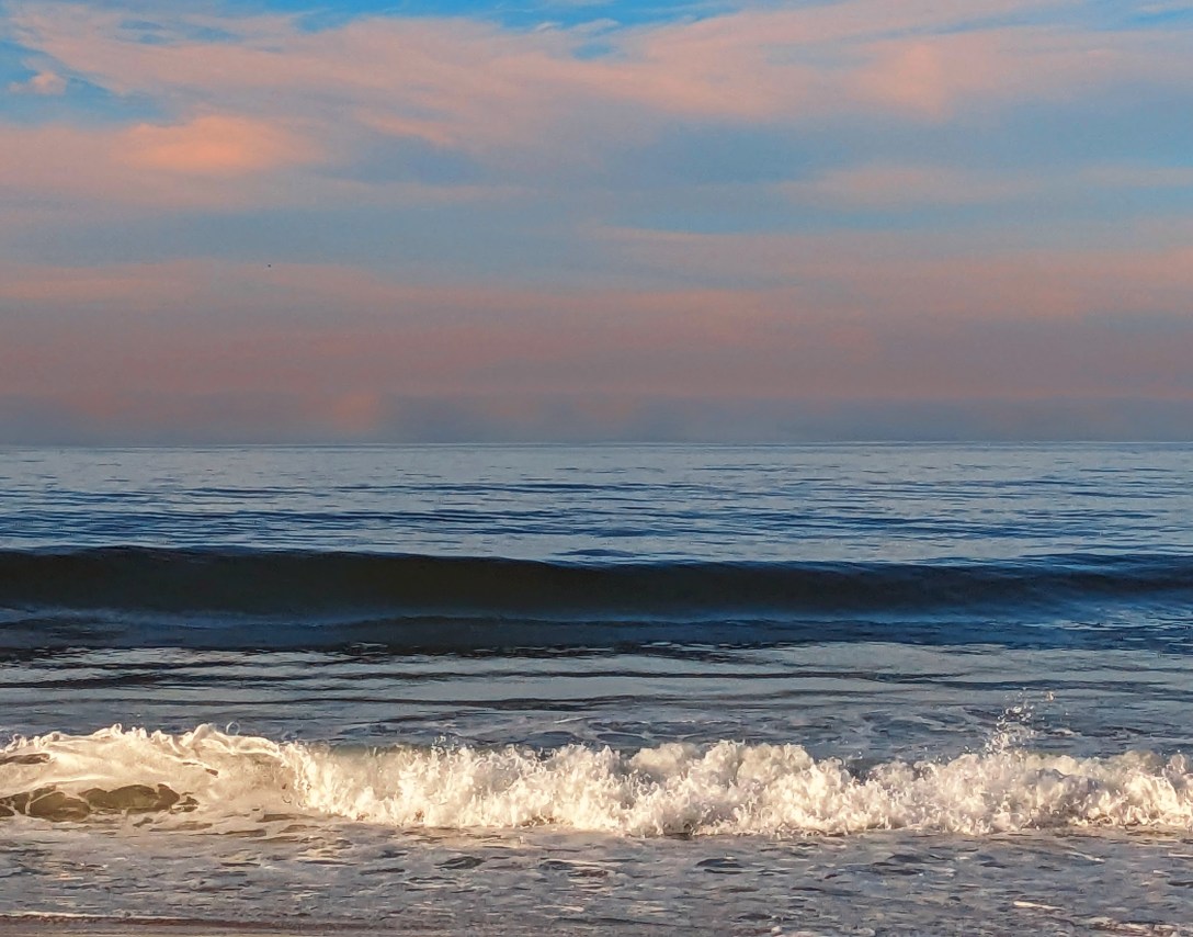 Soft pink and blue morning sky over a calm ocean, with a wave building and breaking into bright white foam near the shore.