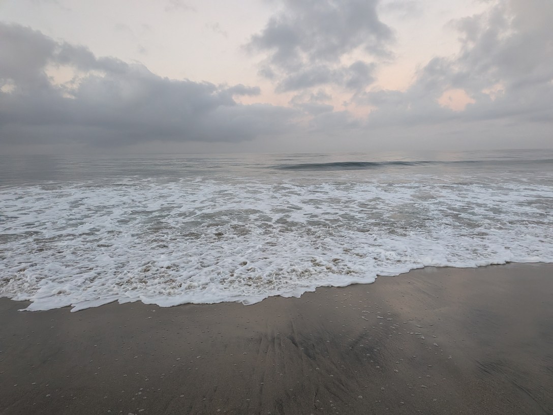 Seafoam spreading over wet sand at dawn, with a small wave forming in the calm water beyond and soft pink light through grey clouds.