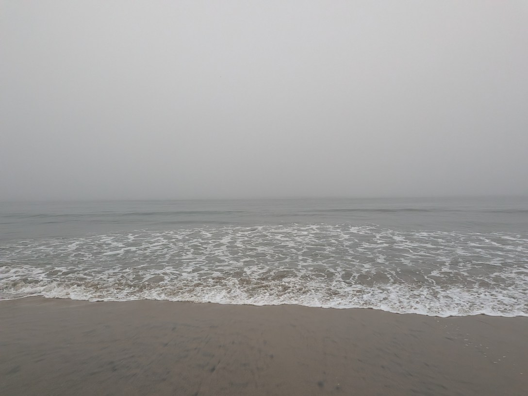 A grey ocean meeting the shore under heavy fog, white foam spreading across wet sand as the horizon fades into the mist.