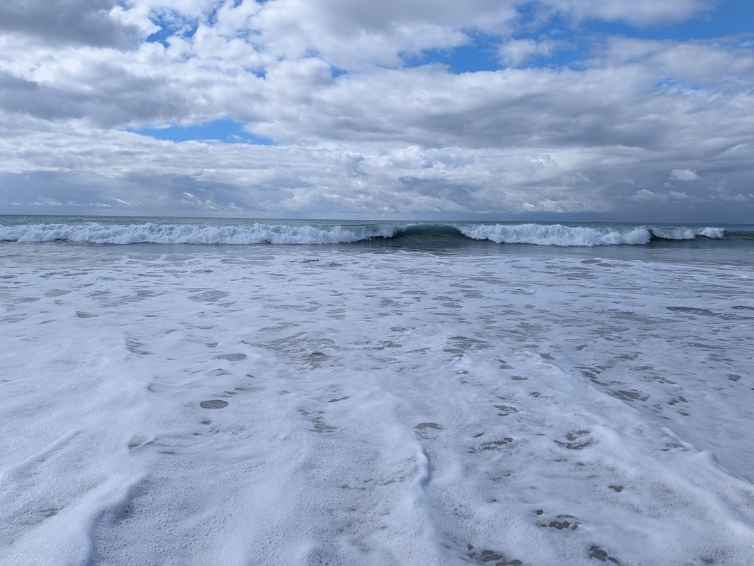 A wave breaks in the distance under a wide, cloudy blue sky while white foam spreads calmly across the sand in the foreground.