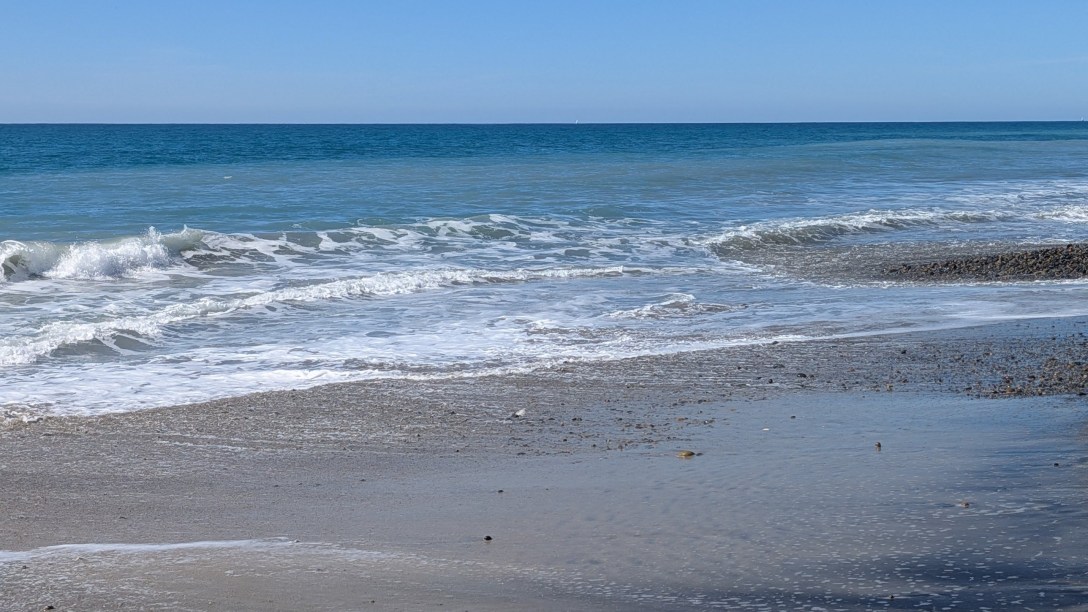 Waves rolling onto a pebbled shoreline under a clear blue sky, a thin sheet of water receding over wet sand.